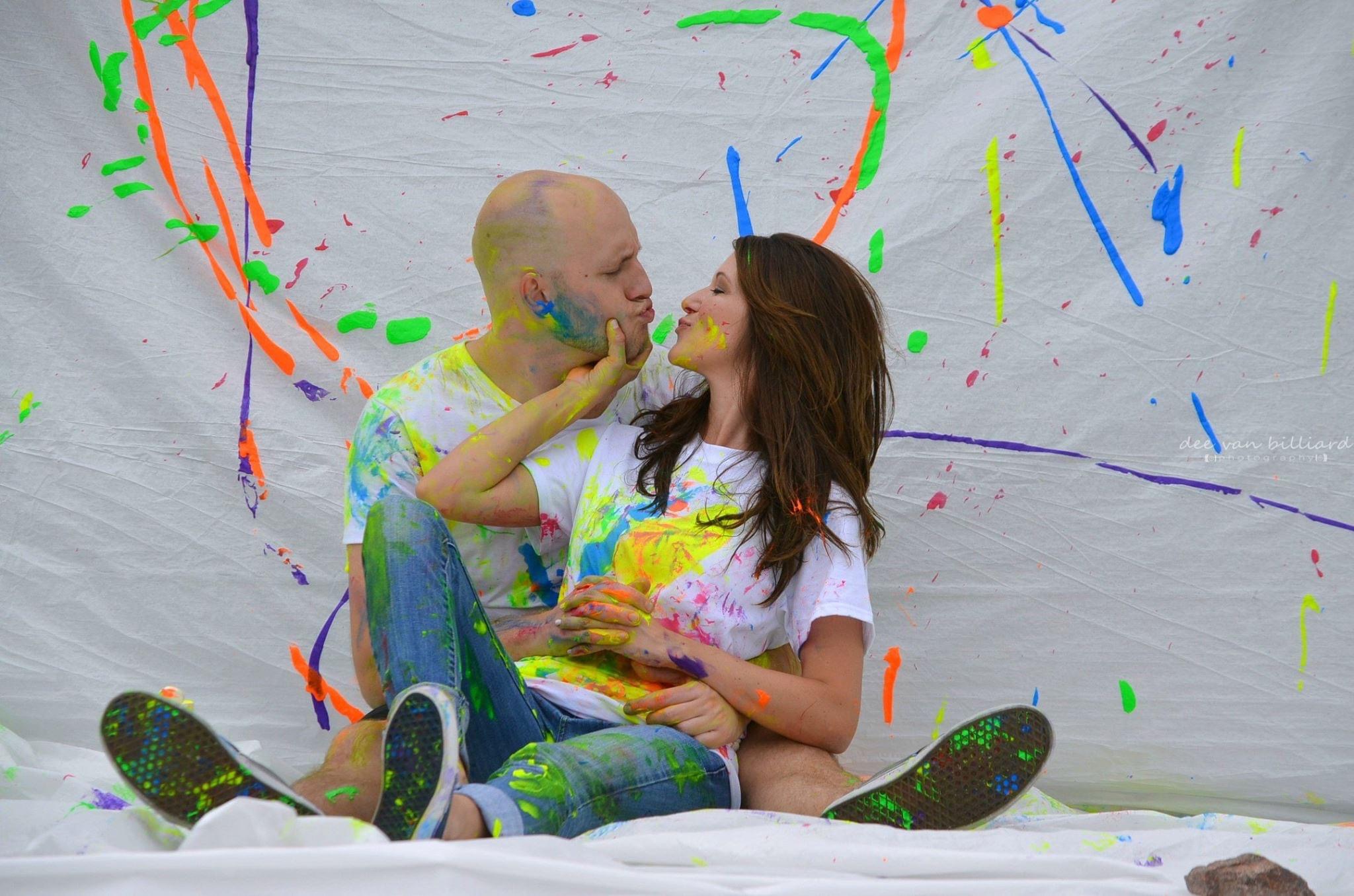 A couple splattered with paint poses in front of a banner.