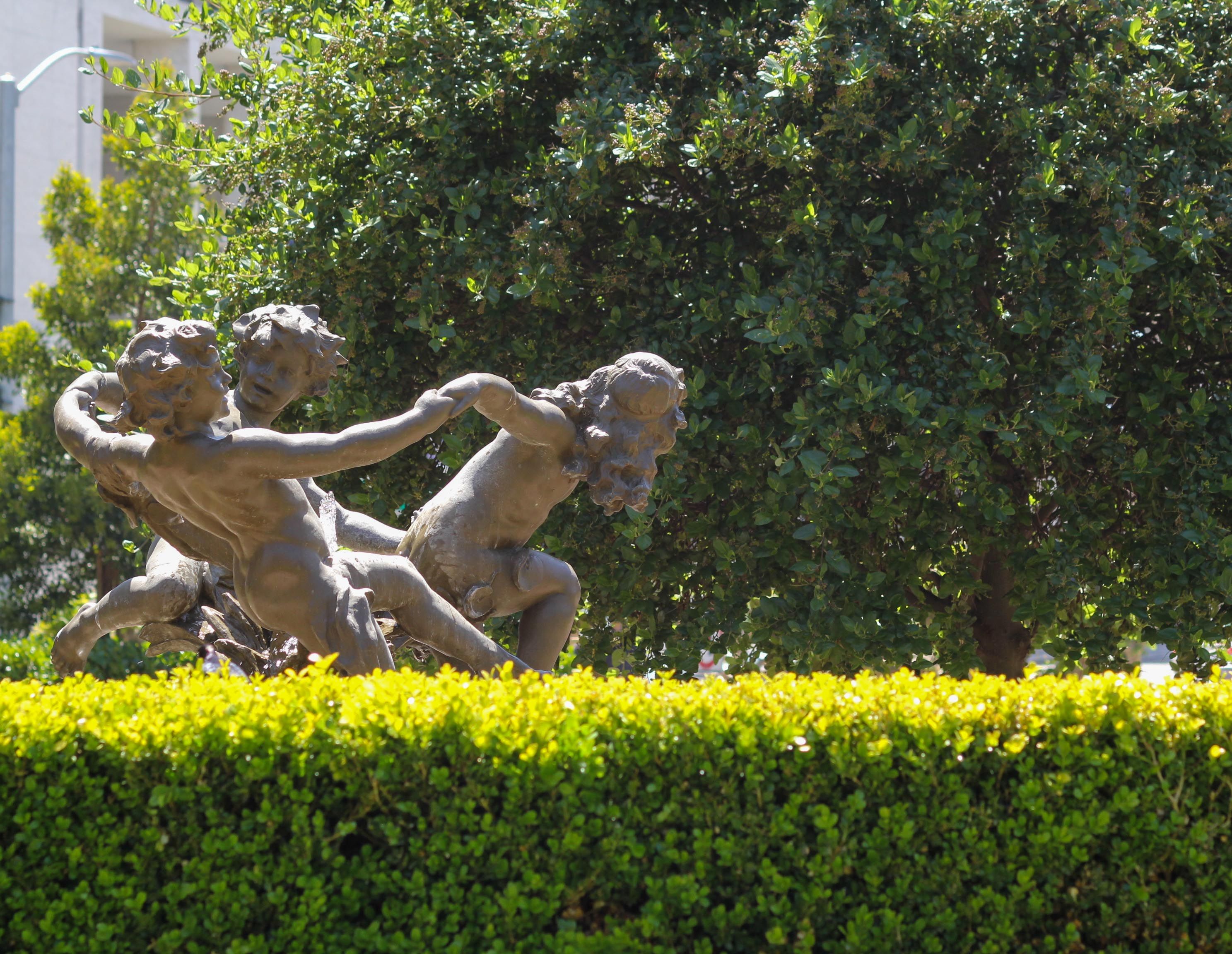 A statue of three people dancing among a wall of tall bushes.