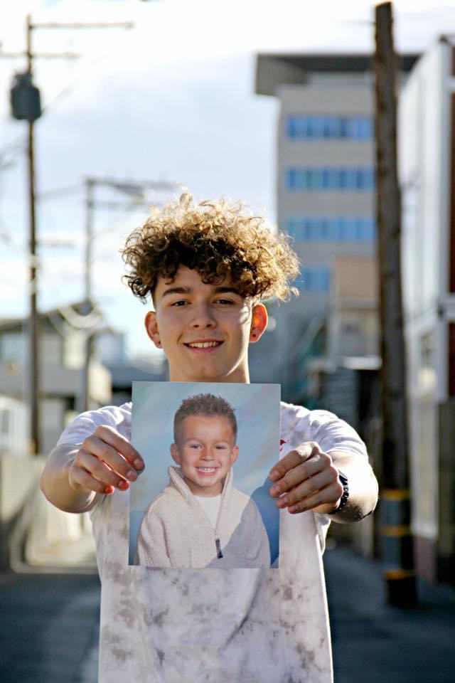A young man with curly hair smiles while holding a baby photo of himself.