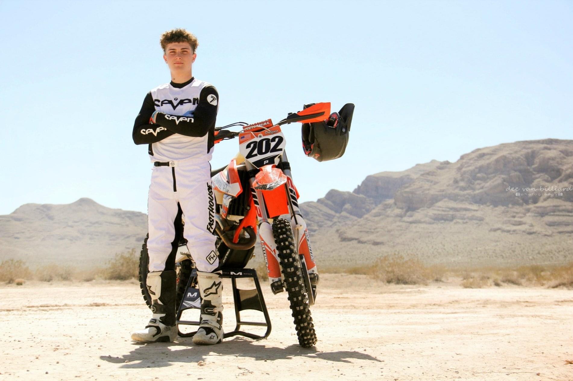A young man stands in front of his dirt bike out in the desert.