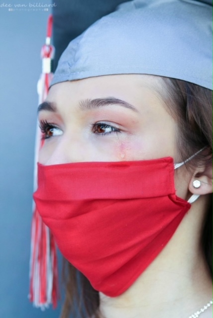 A close up of a young lady gently crying while wearing a medical mask and graduate cap.