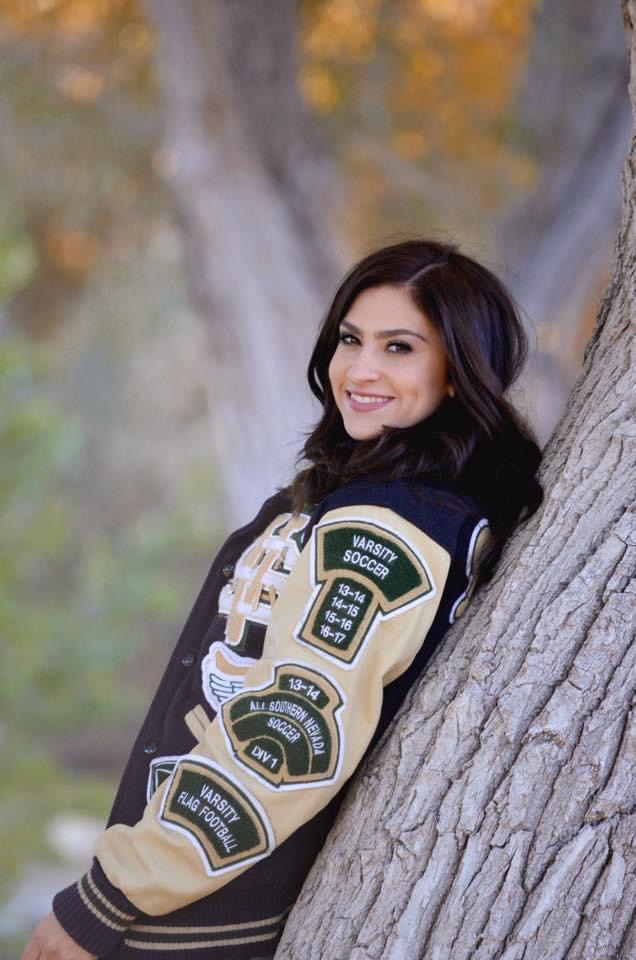 A young lady in a large, varsity jacket smiles while leaning on a tree.