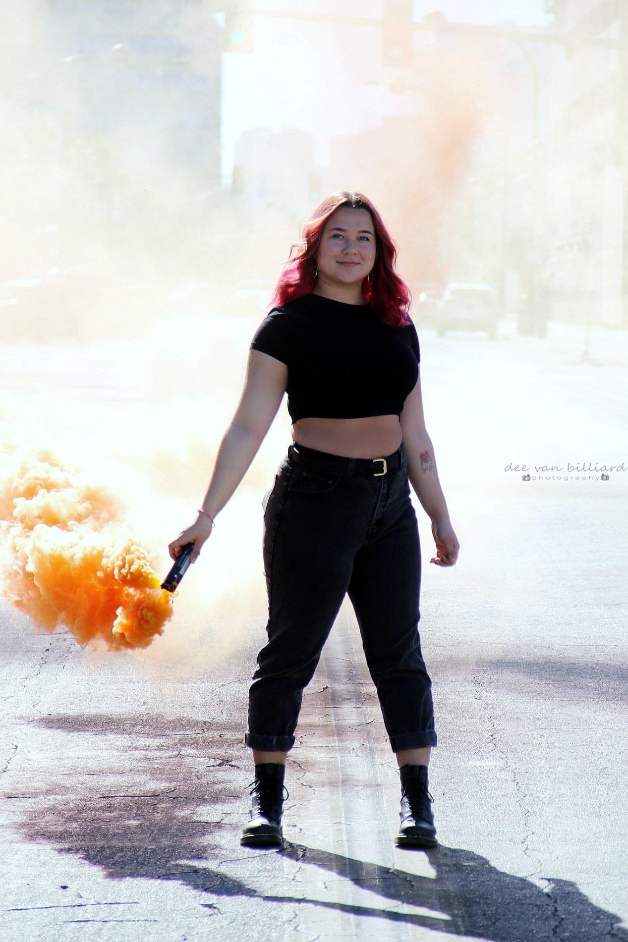 A young lady poses on the street while carrying a stick producing colorful smoke.