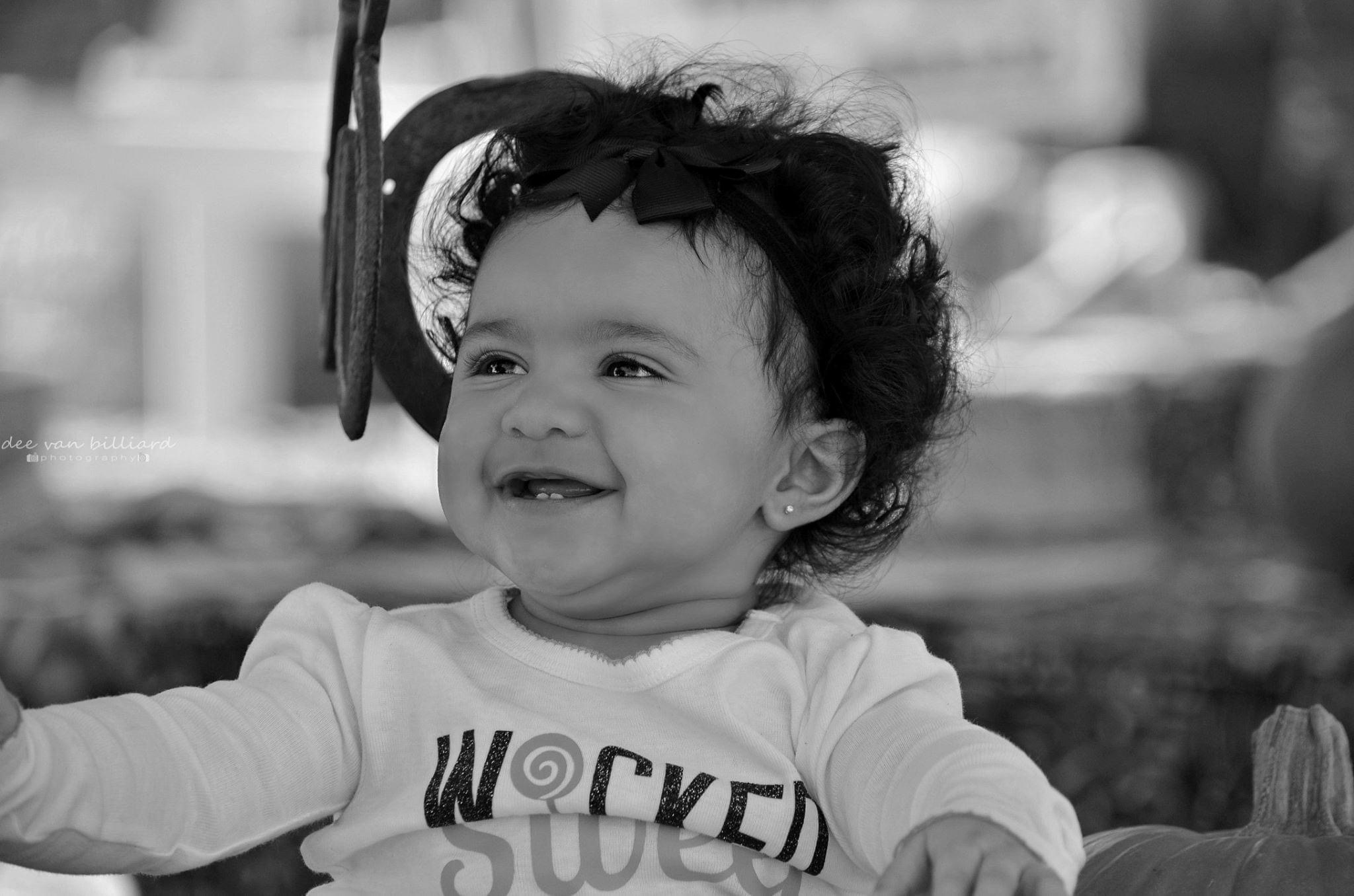 A black and white photo of a baby smiling with a bow on her head.