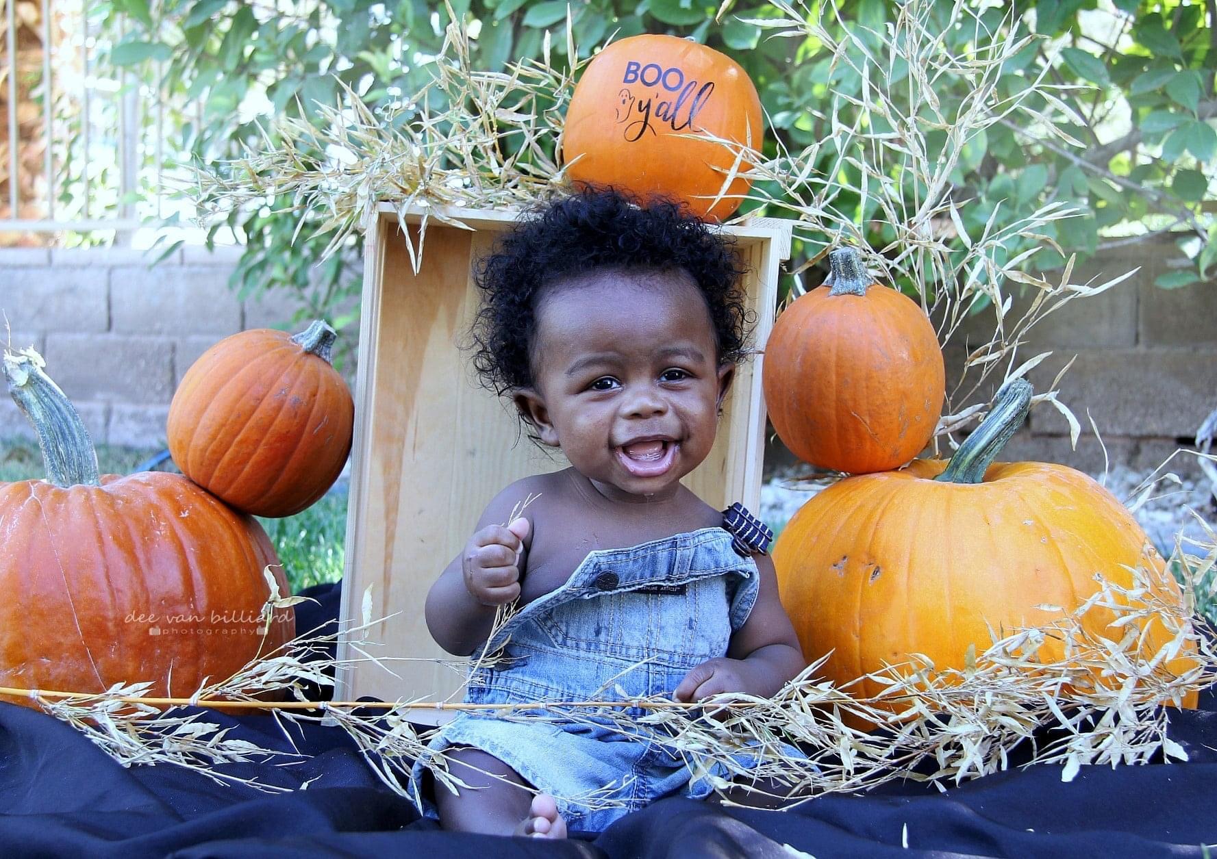 A baby smiling atop a table, sitting among bushel of hay and a few pumpkins.
