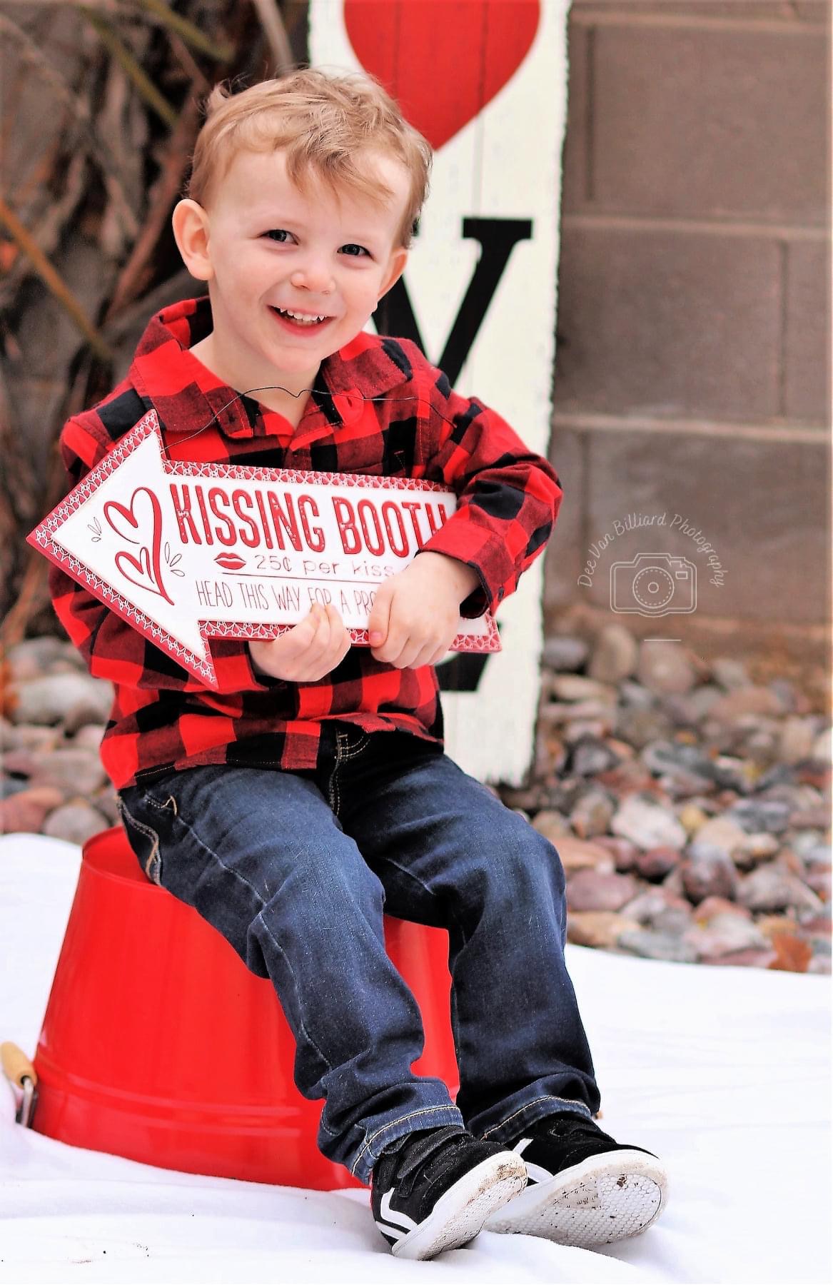 A male toddler smiling while carrying a kissing booth sign.