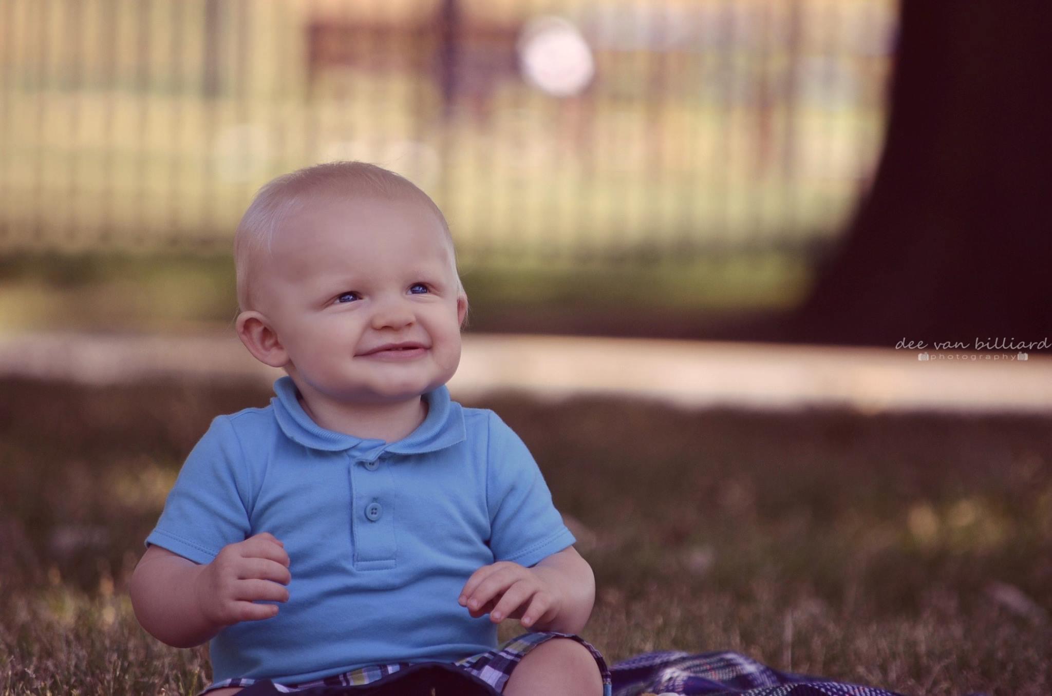 A baby boy sitting at the park, smiling upward.
