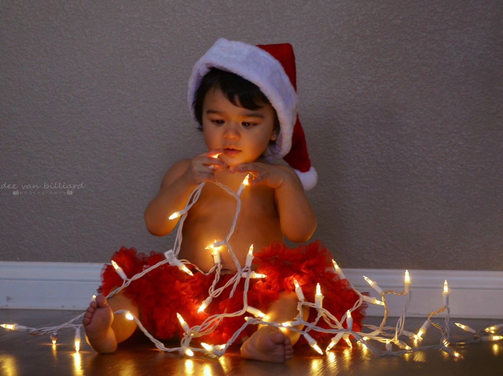 A baby girl in a santa hat sits in a web of Christmas lights.