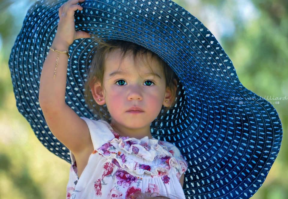 A baby girl in a large floral dress and a large hat looks at the camera.