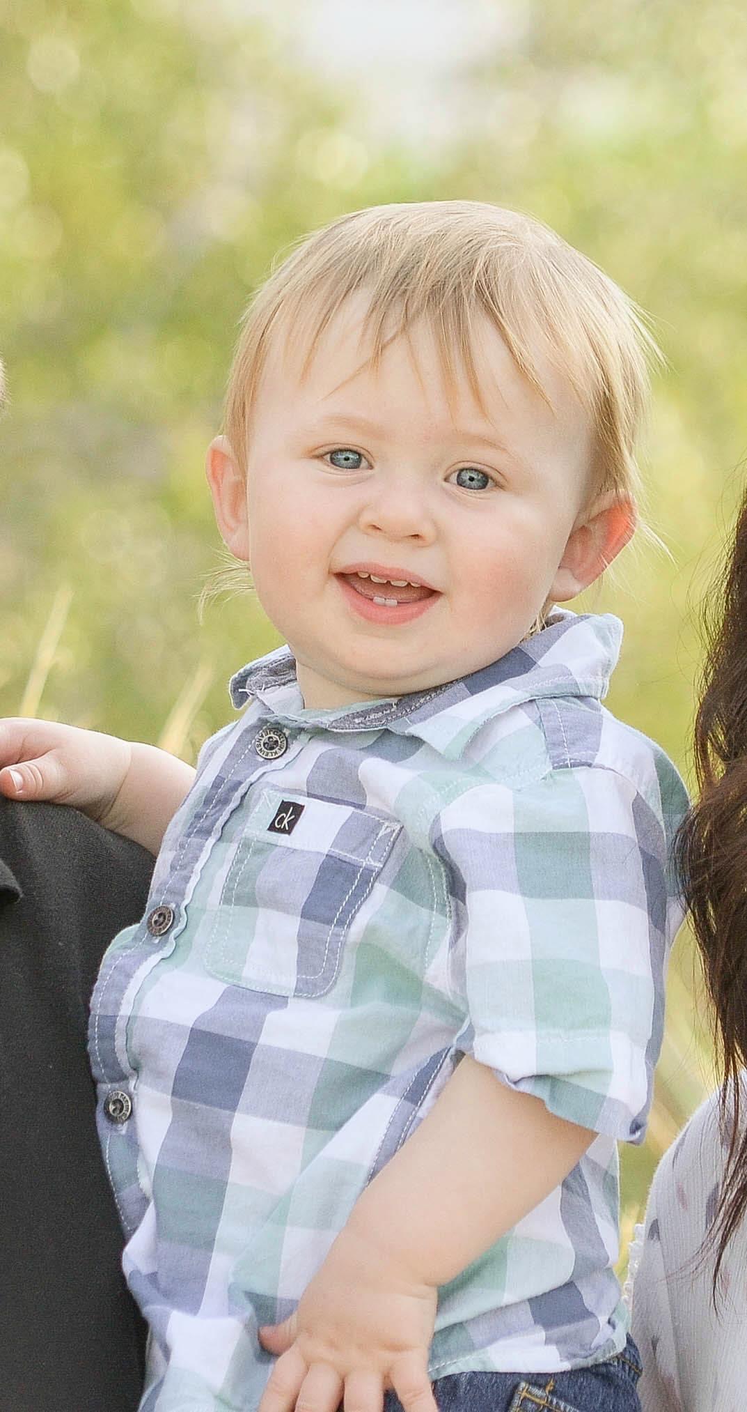 A baby boy with his parents in the grass smiles at the camera.